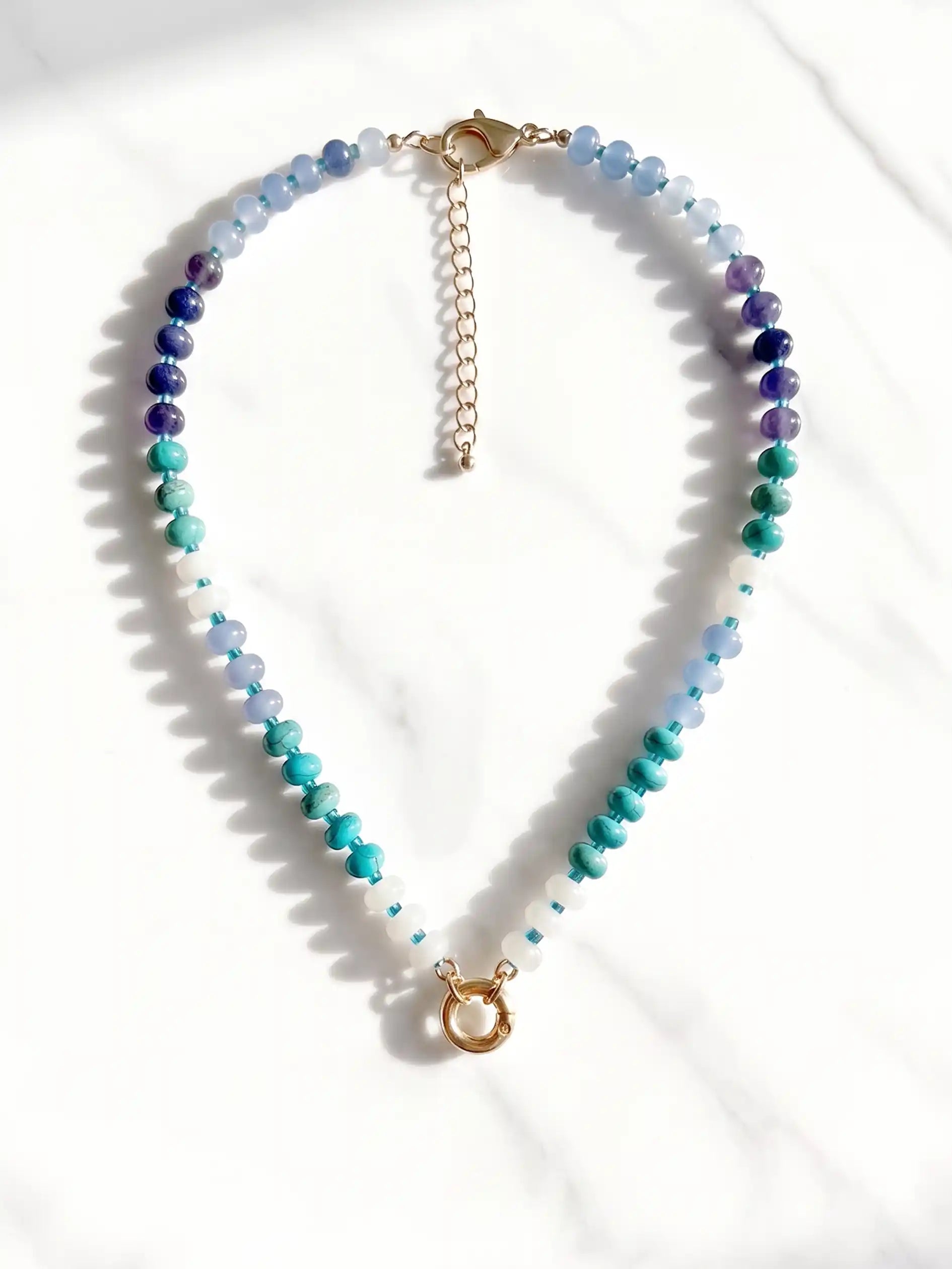 A semi-precious stone beaded necklace with faceted round beads transitioning through shades of blue, teal, green, and purple, accented by small white beads and gold-tone circle carabiner, displayed on a white marble surface.