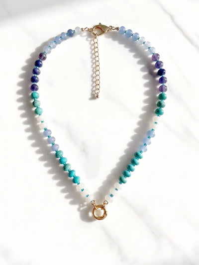 A semi-precious stone beaded necklace with faceted round beads transitioning through shades of blue, teal, green, and purple, accented by small white beads and gold-tone circle carabiner, displayed on a white marble surface.