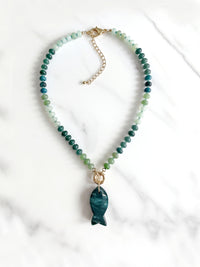A beaded necklace featuring green and blue gradient round beads with a polished dark moss green agate fish-shaped pendant, suspended from a gold ring on a white background.