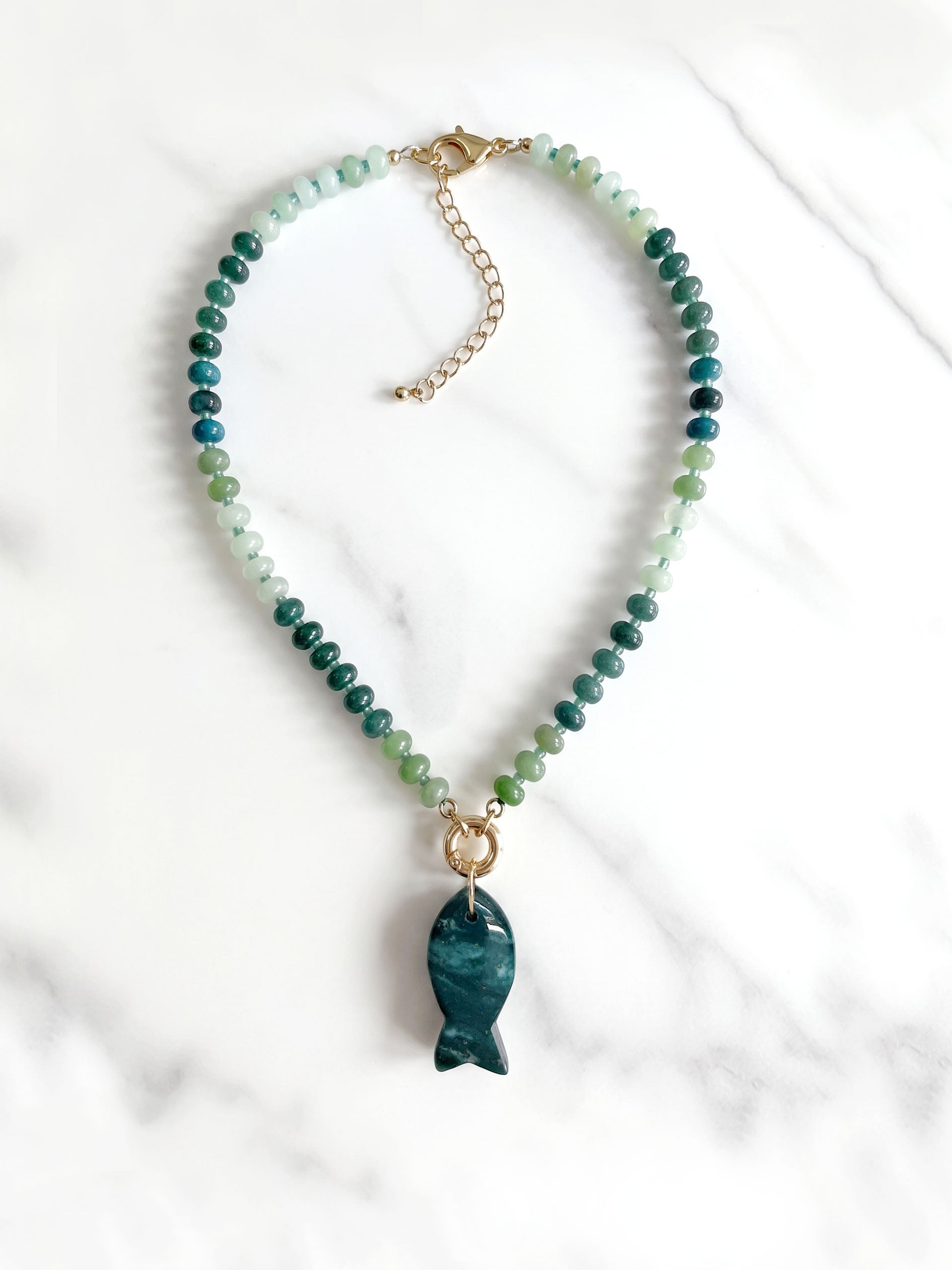 A beaded necklace featuring green and blue gradient round beads with a polished dark moss green agate fish-shaped pendant, suspended from a gold ring on a white background.
