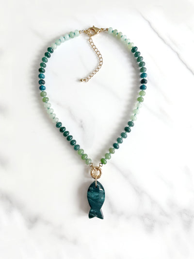 A beaded necklace featuring green and blue gradient round beads with a polished dark moss green agate fish-shaped pendant, suspended from a gold ring on a white background.