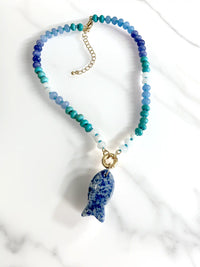 A beaded necklace with faceted beads in shades of blue, teal, and white, featuring a blue sodalite stone fish pendant with gold-tone hardware, displayed on a white marble surface.