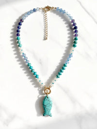 A beaded necklace with faceted blue, teal, and purple beads, a gold-tone chain extender, and a turquoise fish-shaped pendant, displayed on a white marble surface.