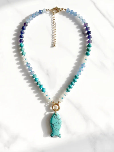 A beaded necklace with faceted blue, teal, and purple beads, a gold-tone chain extender, and a turquoise fish-shaped pendant, displayed on a white marble surface.