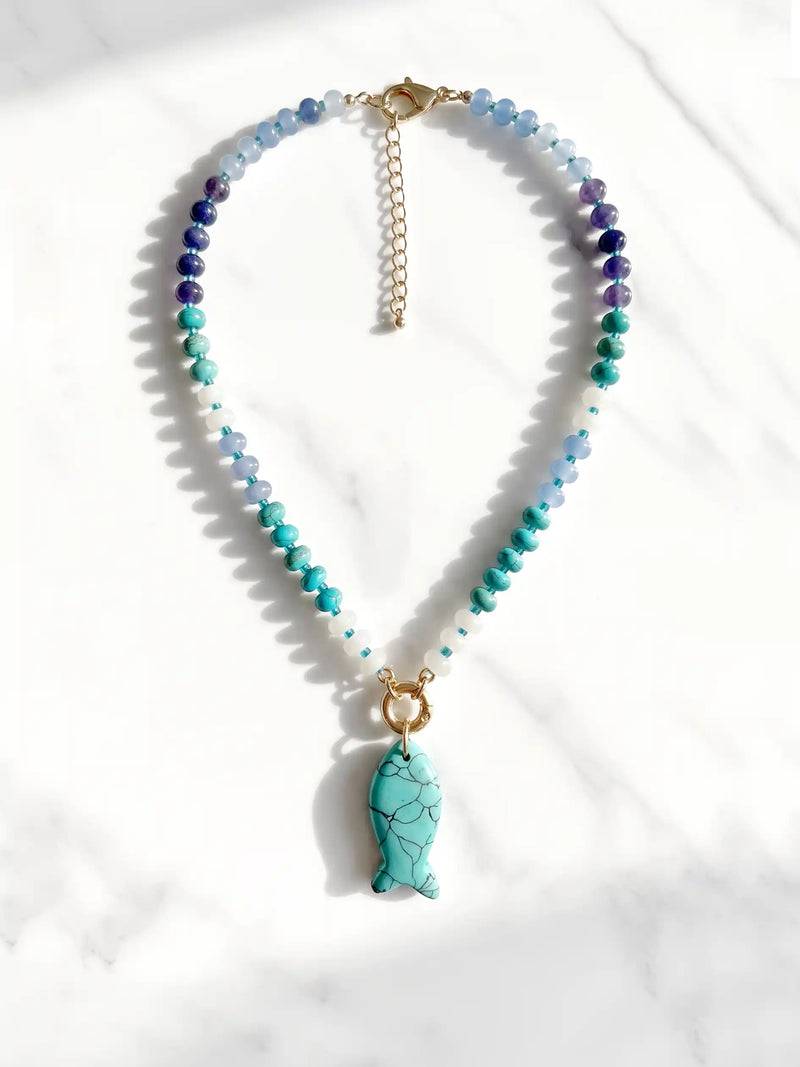 A beaded necklace with faceted blue, teal, and purple beads, a gold-tone chain extender, and a turquoise fish-shaped pendant, displayed on a white marble surface.