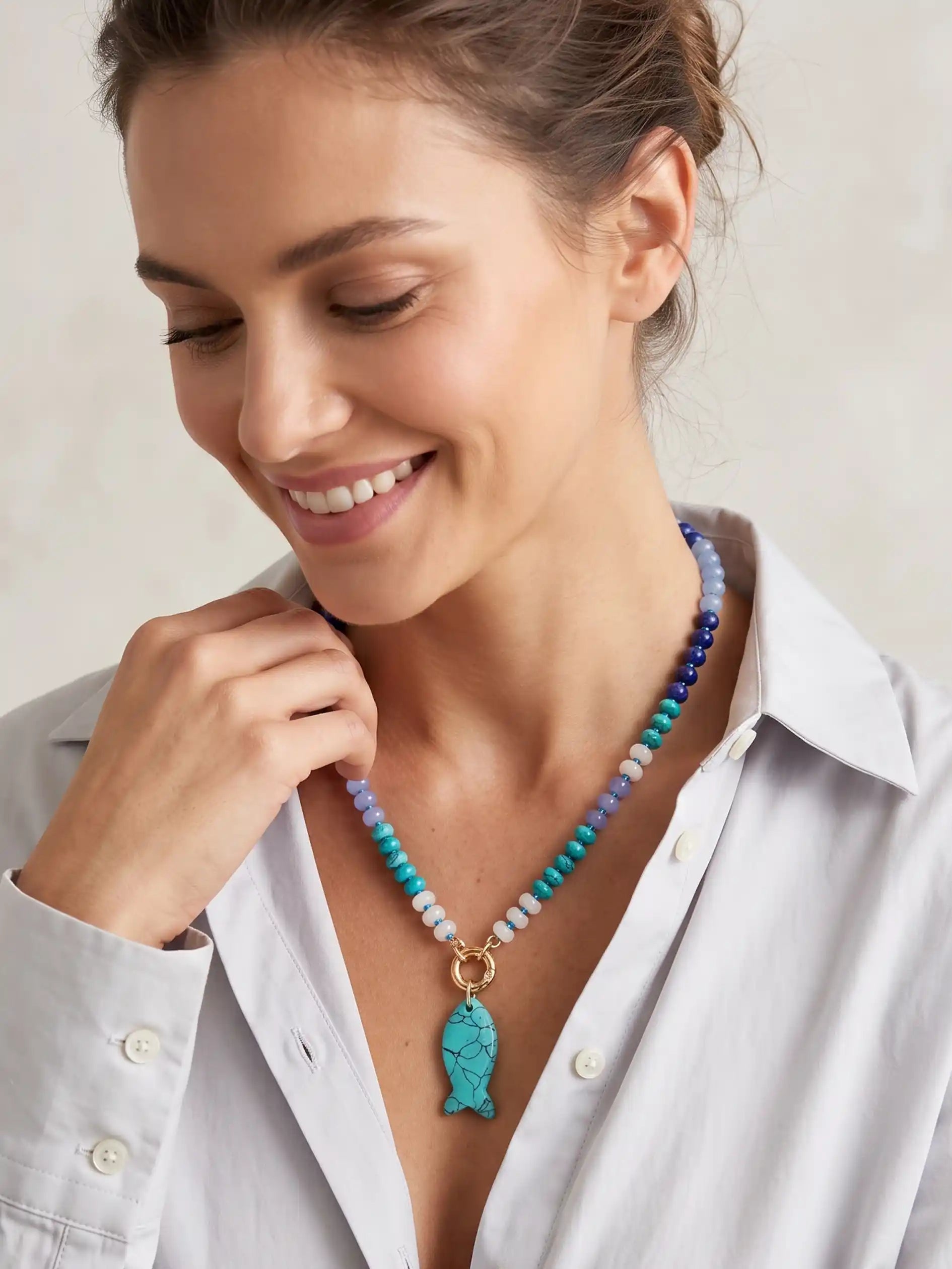 A woman wearing a colorful beaded necklace with a turquoise fish pendant, paired with a white button-down shirt.