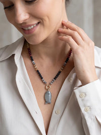 A close-up lifestyle shot of a woman wearing a beaded necklace with a gray stone pendant. The necklace features multi-colored beads including black, gray, white, and red, accented by gold-toned metal rings.