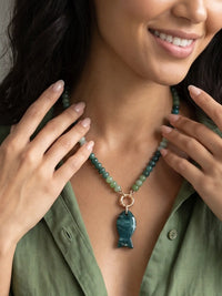 A close-up lifestyle shot of a woman wearing a beaded necklace with a polished green fish pendant and rose gold clasp, paired with an olive green shirt.