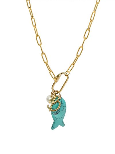 A gold paperclip chain necklace featuring a front-facing carabiner clasp that holds three distinct charms: a small round pearl, a gold script initial B, and a carved turquoise blue fish pendant.