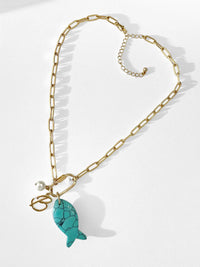A gold paperclip chain necklace featuring a front-facing carabiner clasp that holds three distinct charms: a small round pearl, a gold script initial B, and a carved turquoise fish pendant.