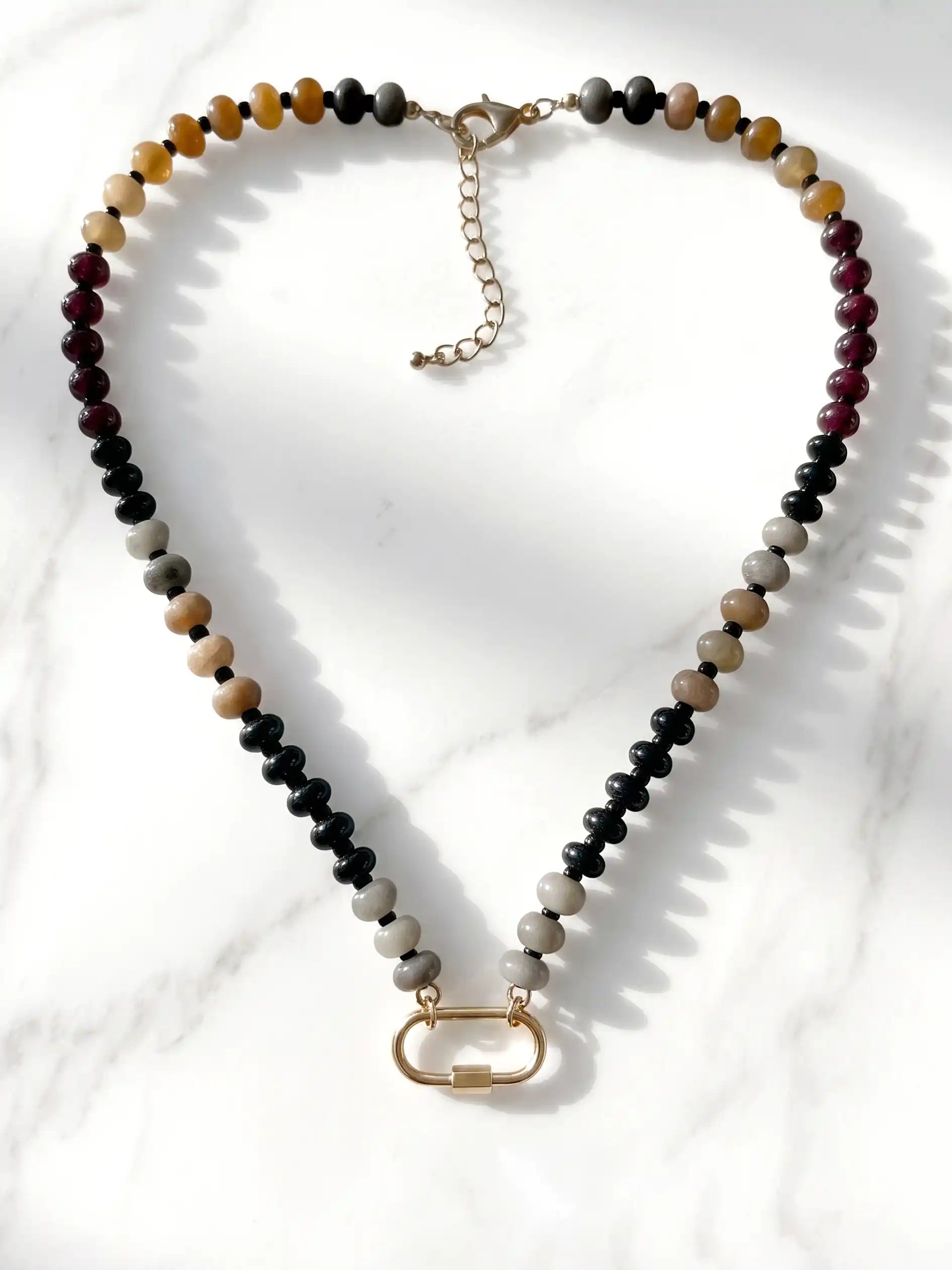 A semi-precious stone beaded necklace with alternating black, gray, tan, and burgundy beads, finished with a gold-tone carabiner clasp and extender chain, displayed on a white marble surface.