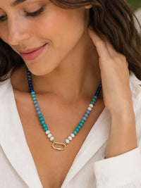 A close-up lifestyle shot of a woman wearing a semi-precious stone beaded necklace with a gold carabiner. The necklace features beads in shades of blue, teal, and white, and is styled with a white collared shirt.