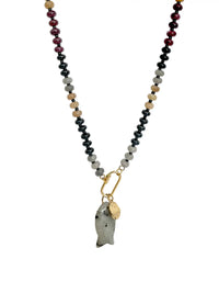 A beaded necklace featuring a central gold carabiner holding a polished gray stone fish pendant and a small gold tag, with the chain extending to the top edge. The chain itself is composed of alternating beads of black, light gray, tan, and deep burgundy on a plain white background.