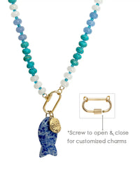 A beaded necklace with a gradient of blue, teal, and white beads, featuring a fish-shaped pendant made of sodalite and a small gold coin charm, displayed on a white marble background with soft leaf shadows.