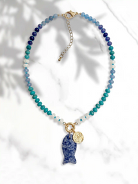 A beaded necklace with a gradient of blue, teal, and white beads, featuring a fish-shaped pendant made of sodalite and a small gold coin charm, displayed on a white marble background with soft leaf shadows.