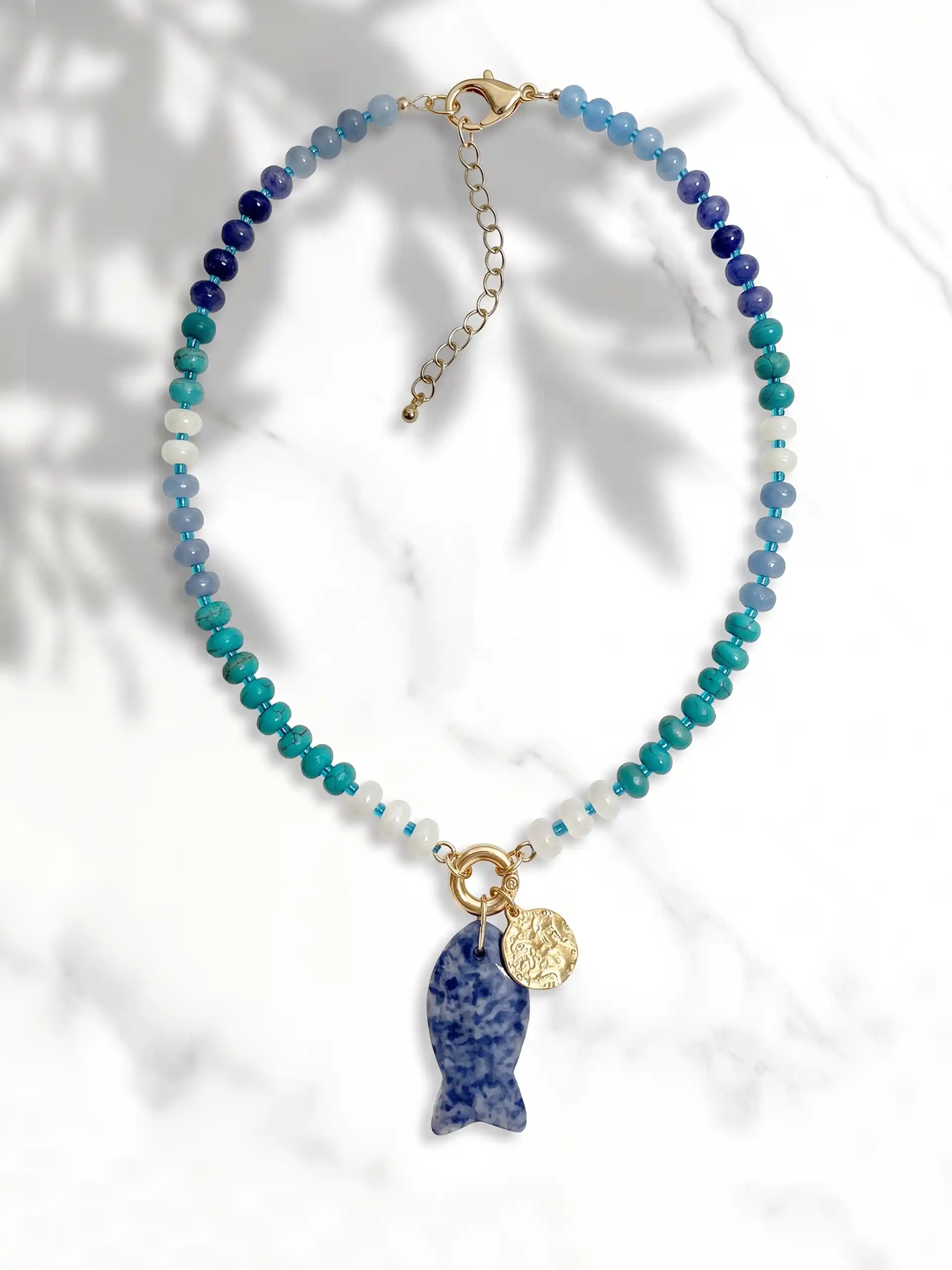 A beaded necklace with a gradient of blue, teal, and white beads, featuring a fish-shaped pendant made of sodalite and a small gold coin charm, displayed on a white marble background with soft leaf shadows.