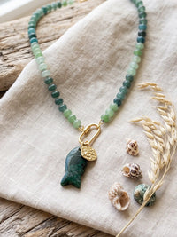 A beaded necklace with green and blue-green round beads, a gold carabiner clasp, and a carved dark green fish pendant, styled on a beige linen cloth with small shells and dried grass.