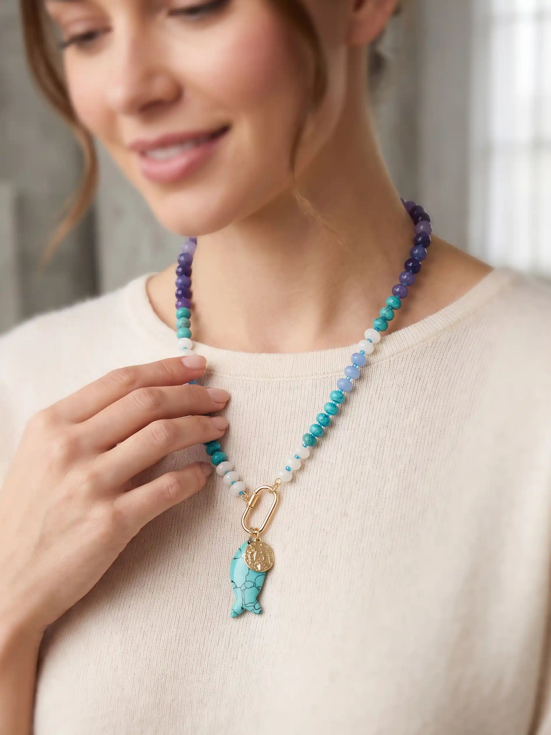 A woman wearing a beaded necklace with turquoise, blue, purple, and white beads, featuring a gold-tone carabiner and a turquoise fish charm, paired with a cream-colored knit top.