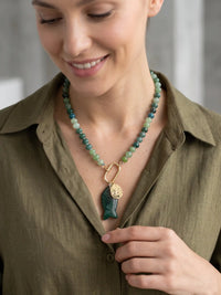 A close-up lifestyle shot of a woman wearing a beaded necklace with a green carved fish pendant and gold charms, paired with an olive green shirt.