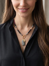 A close-up of a woman wearing a beaded necklace with a gray stone pendant and gold-toned carabiner, paired with a black blouse.