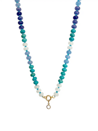 A beaded necklace featuring alternating strands of white, light blue, teal, and dark blue beads, finished with a gold ring with a small crystal stone point charm on a solid white background.