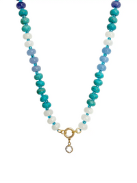A top-down photograph of a unique handmade semi-precious stone beaded necklace against a white background. The necklace features alternating deep blue, turquoise, light blue, and white stone beads. At the center is a gold-colored round ring with a small, clear crystal point pendant hanging from it.