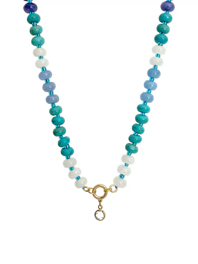 A top-down photograph of a unique handmade semi-precious stone beaded necklace against a white background. The necklace features alternating deep blue, turquoise, light blue, and white stone beads. At the center is a gold-colored round ring with a small, clear crystal point pendant hanging from it.