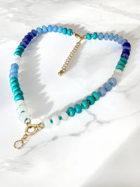 A heart-shaped beaded necklace with a gradient of blue, teal, and white beads, accented by gold-tone ring, displayed on a white marble surface.