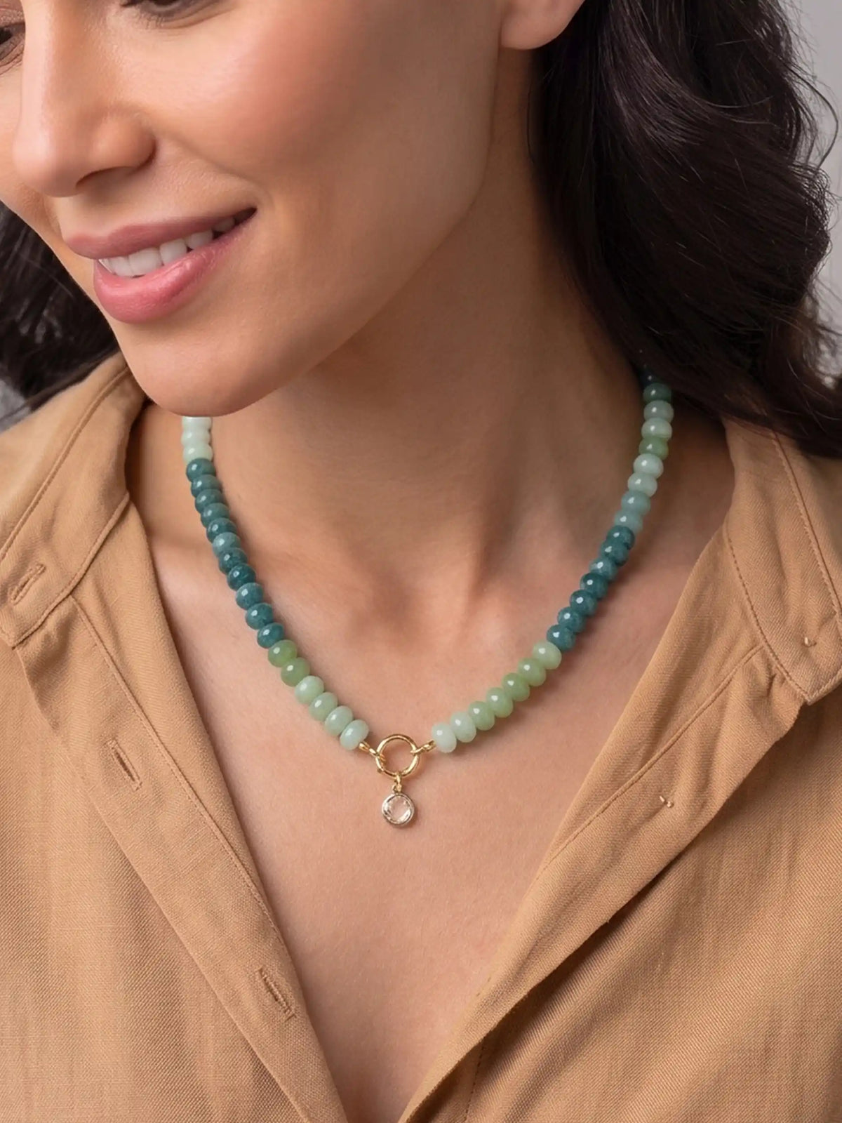A close-up lifestyle shot of a woman wearing a green tone beaded necklace with a gold metal ring and a small clear stone pendant