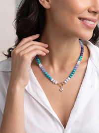 A close-up of a woman wearing a beaded necklace with turquoise, light blue, and purple beads, accented by a gold ring and a small clear stone pendant.
