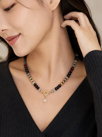 A close-up lifestyle shot of a woman wearing a beaded necklace with a gold metal ring and a small clear stone pendant, paired with a black ribbed top.