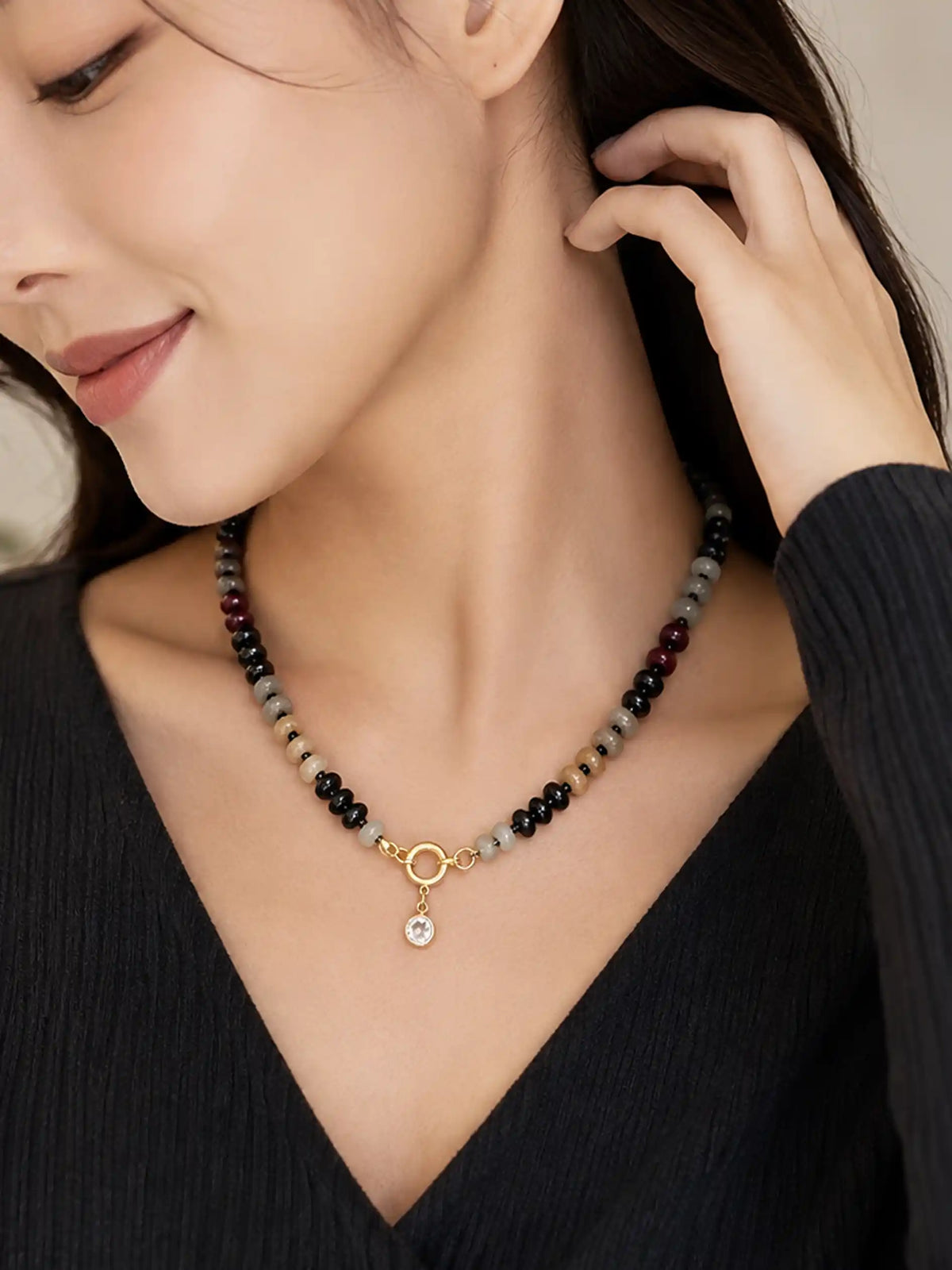 A close-up lifestyle shot of a woman wearing a beaded necklace with a gold metal ring and a small clear stone pendant, paired with a black ribbed top.