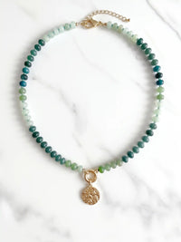 Green beaded statement necklace with mixed green beads, gold-tone clasp, and textured round coin pendant on a white marble stone background.