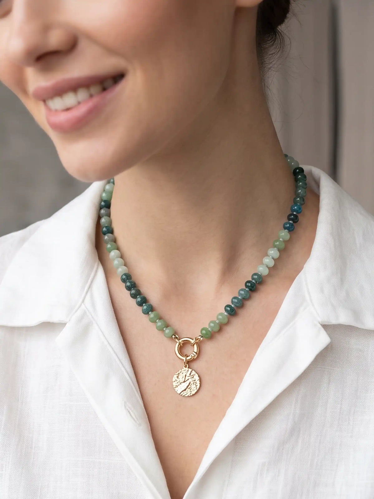 Green beaded statement necklace with mixed green beads, gold-tone clasp, and textured round coin pendant on a white background.