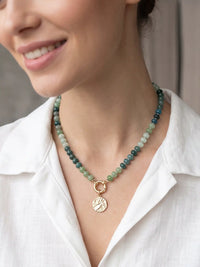 Green beaded statement necklace with mixed green beads, gold-tone clasp, and textured round coin pendant on a white background.
