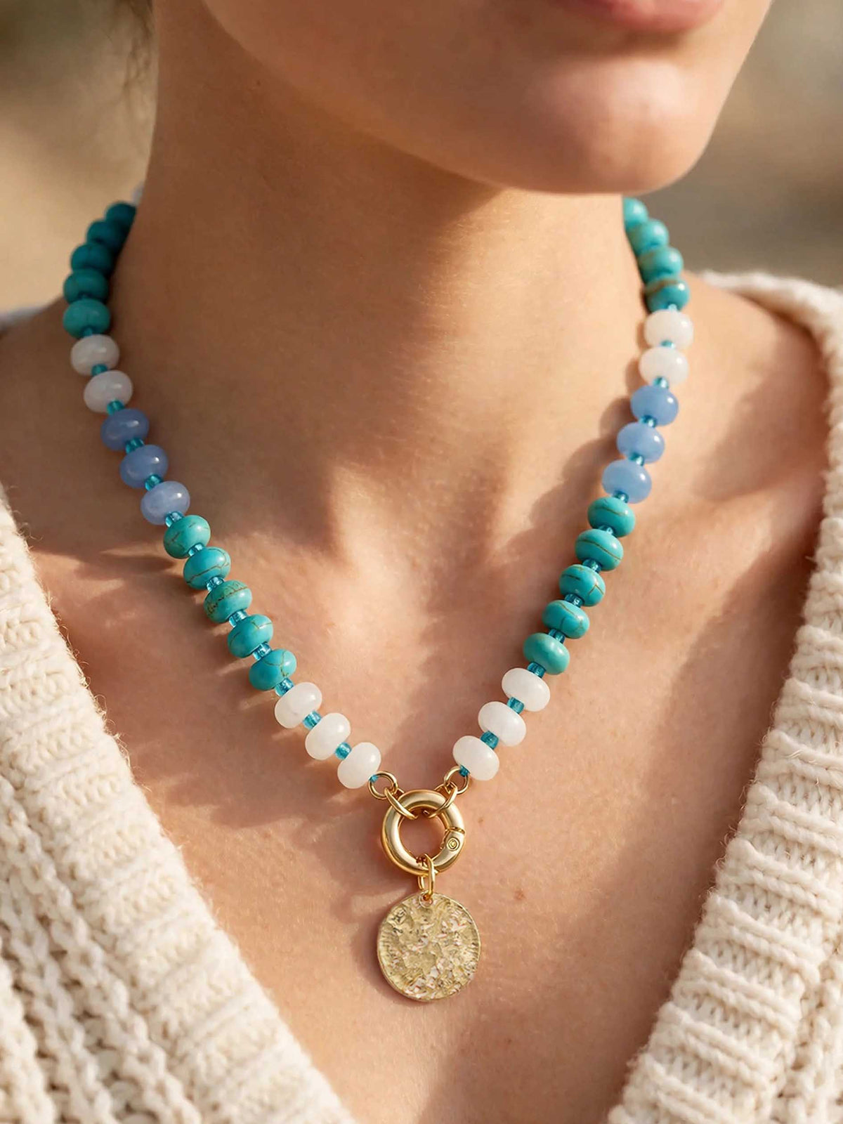 A close-up of a model wearing a beaded necklace with sections of turquoise, opaque white, and translucent blue beads. The necklace features a front-facing gold carabiner clasp holding a textured gold circular pendant. The model is wearing a cream-colored chunky knit sweater.