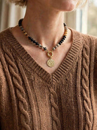 A close-up of a model wearing a beaded necklace featuring sections of black, grey, and amber-brown beads. The necklace has a front-facing gold carabiner clasp holding a textured gold circular pendant. The model is wearing a brown cable-knit V-neck sweater.