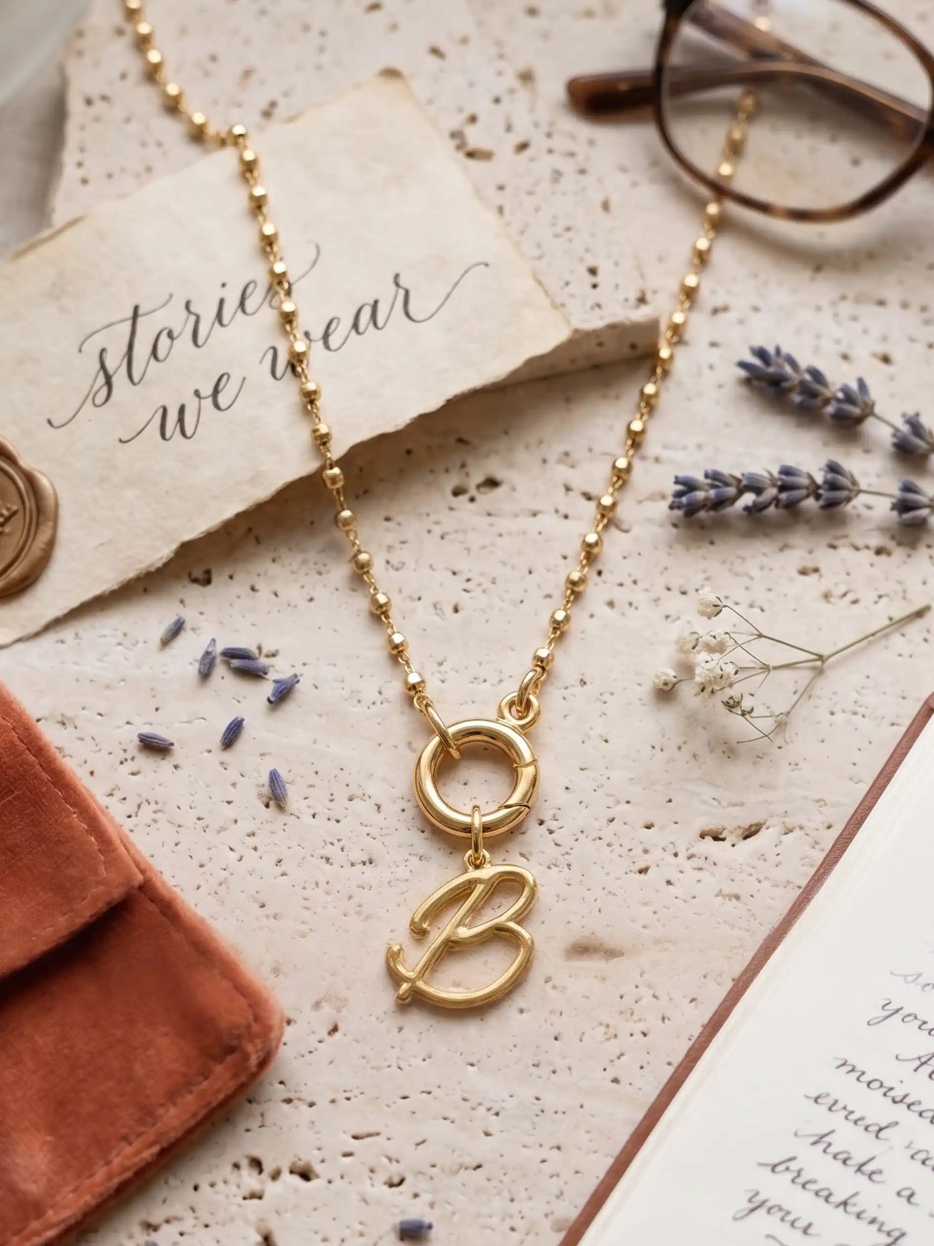 A flat-lay photograph on a textured stone surface, featuring a central gold necklace with a script initial 'B' pendant. Arranged around it are a pair of tortoiseshell glasses, an open leather notebook with handwriting, dried lavender and baby's breath, and an aged paper tag reading 'stories we wear' with a wax seal.