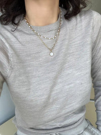 A close-up of a person wearing a light gray ribbed long-sleeve top and layered necklaces, including one with small white pearls and one with a round white pendant on a gold chain.