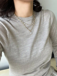 A close-up of a person wearing a light gray ribbed long-sleeve top and layered necklaces, including one with white pearls and one with a small round pendant.