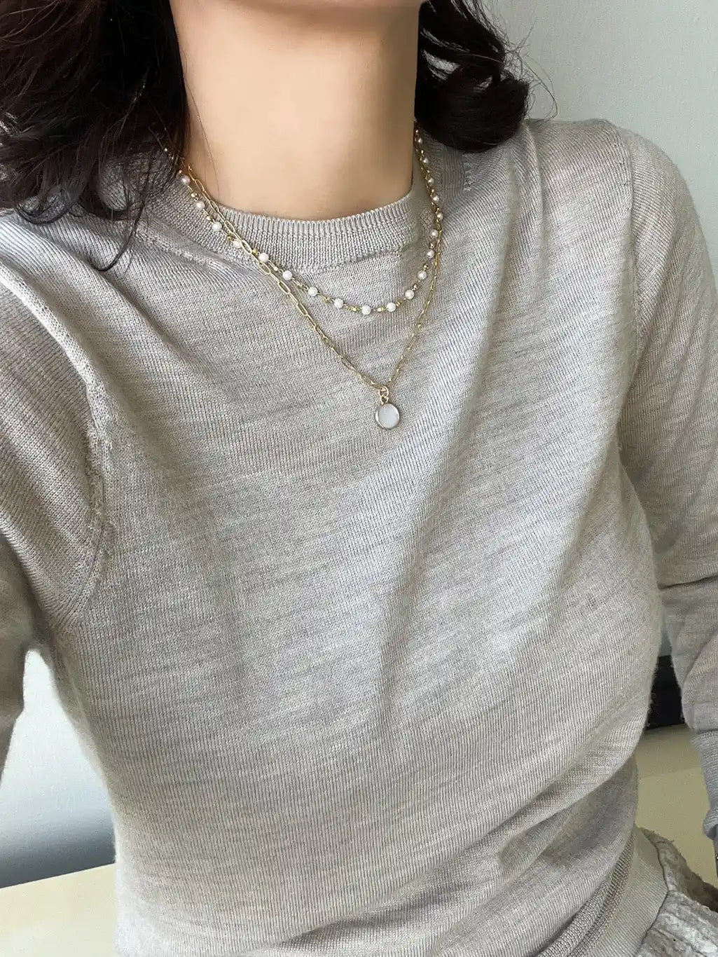 A close-up of a person wearing a light gray ribbed long-sleeve top and layered necklaces, including one with white pearls and one with a small round pendant.