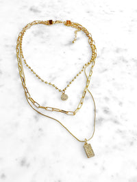 A layered gold necklace set featuring a paperclip chain, a beaded ball chain with a small round pavé charm, and a delicate chain with a rectangular pavé pendant, displayed on a white marble background.