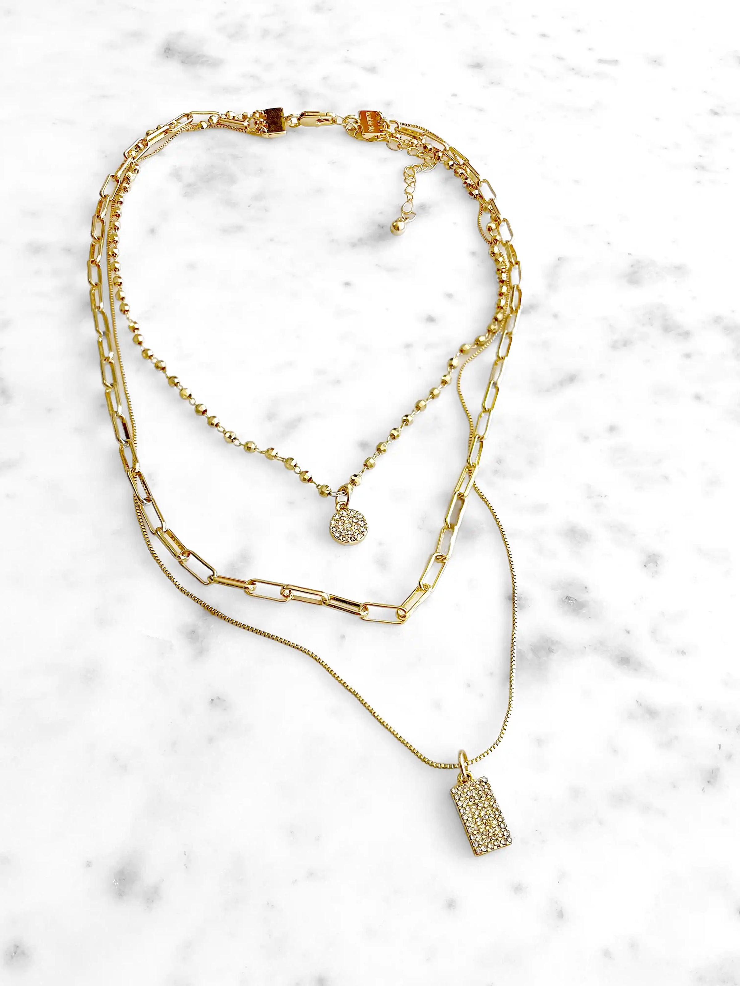 A layered gold necklace set featuring a paperclip chain, a beaded ball chain with a small round pavé charm, and a delicate chain with a rectangular pavé pendant, displayed on a white marble background.