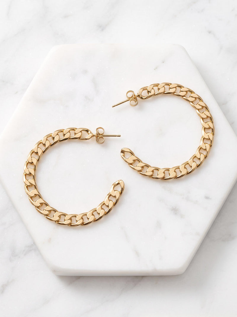 Gold chain-link hoop earrings displayed on a white hexagonal marble tray over a light marble surface.