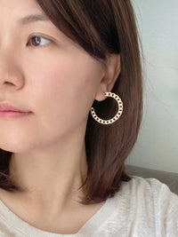 A close-up of a woman wearing a large gold hoop earring with small black dot accents, set against a soft, neutral indoor background.