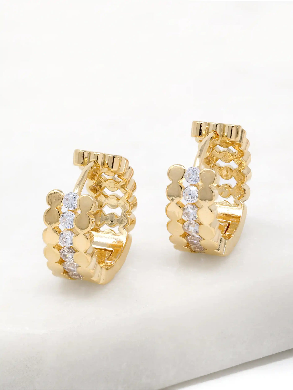 A pair of gold huggie hoop earrings with a textured, layered design featuring small gold circles and clear pavé-set round stones, displayed on a white surface.