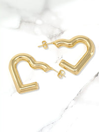 A pair of gold-toned, wavy open heart-shaped hoop earrings with post-back closures, displayed on a white marble surface.