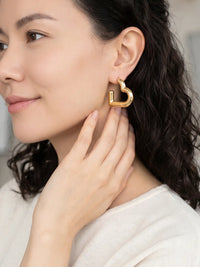 A close-up of a woman wearing a polished gold geometric hoop earring, touching her neck, with her dark hair and white top visible against a soft, blurred background.