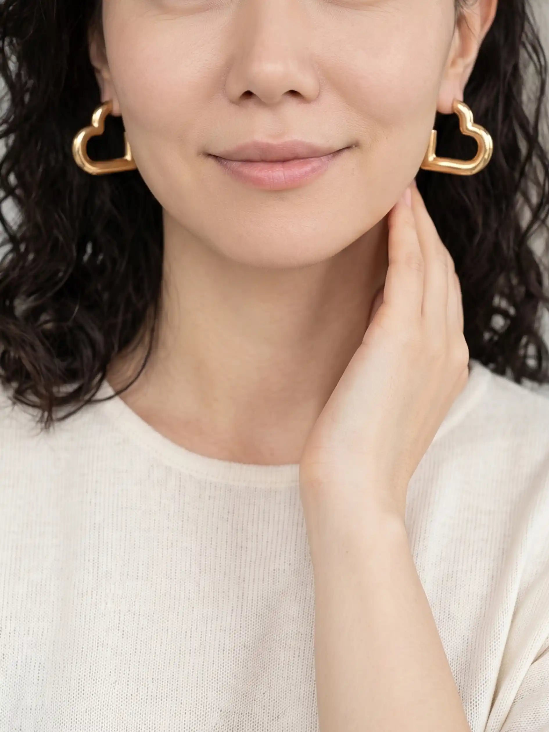 A close-up of a woman wearing large open heart-shaped gold earrings and a cream-colored knit top.