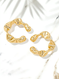 A pair of gold-toned abstract wavy hoop cuff earrings displayed on a white marble surface with soft palm leaf shadows.